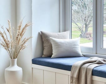 May include: A tall white ceramic vase filled with dried beige pampas grass stems. A window seat features a dark blue cushion, a square beige textured pillow, a rectangular white textured pillow, and a beige fringed throw blanket draped over the edge.