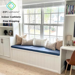 May include: A white window seat with a blue cushion and several throw pillows. The seat is built into a white wall with white trim. The window has a white blind and looks out onto a green yard.