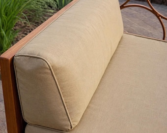 May include: Close-up of a section of an outdoor sofa. The sofa features a wooden frame and tan cushions with a light-colored piping. The cushions are rectangular and appear comfortable. The background shows greenery and a decorative metal railing.