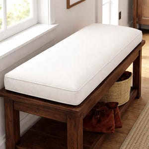 May include: A wooden bench with a white cushion and a dark brown frame. The bench has a lower shelf with a woven basket and two brown drawstring bags. The bench is near a window.