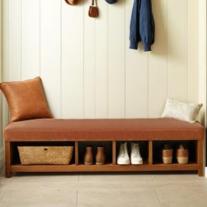 May include: A brown leather upholstered bench with a storage shelf underneath. The bench has three compartments for storing shoes. The bench is in front of a white paneled wall.