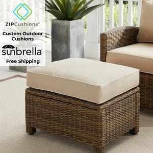 May include: A brown wicker ottoman with a beige cushion. The ottoman is sitting on a beige rug. The text "ZIPCushions® Custom Outdoor Cushions sunbrella Free Shipping" is visible in the image.