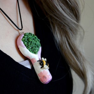 May include: A handmade pendant necklace featuring a pink, white, and purple pipe design with a green textured filling. A small bee and pink flower accents the pipe. The pendant hangs from a black cord.