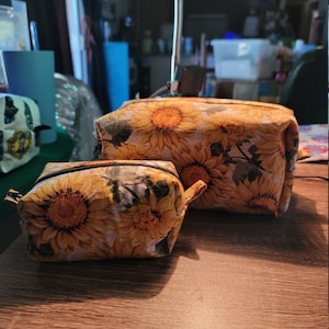 May include: Two zippered pouches with a sunflower print. The larger pouch is brown and the smaller pouch is yellow.