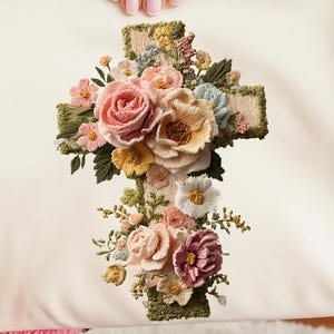 May include: Cream-colored pillow with an embroidered cross design. The cross is adorned with a variety of colorful flowers, including pink, peach, and yellow roses, and other floral accents. The pillow is held by a person.