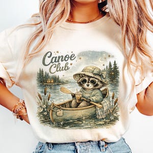Könnte beinhalten: Cremefarbenes T-Shirt mit einem Waschbären in einem Kanu auf einem See. Der Waschbär trägt eine Sonnenbrille und einen Hut und hält ein Paddel und ein Getränk. Die Worte "Canoe Club" stehen über dem Waschbären. Der Hintergrund zeigt Bäume, Wasser und einen Sternenhimmel.