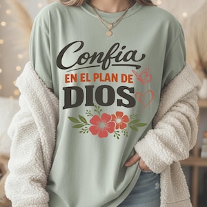 May include: Sage green t-shirt with the Spanish text "Confia en el plan de Dios" in black and orange. The design includes red flowers, green leaves, a butterfly, and a heart. The shirt is worn with a cream-colored, fuzzy jacket.