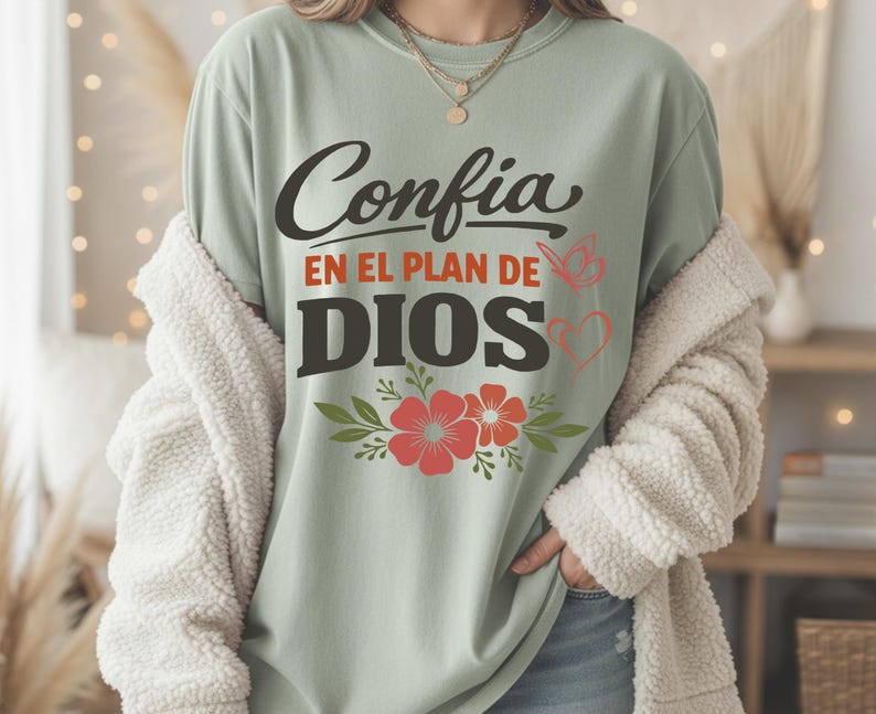 May include: Sage green t-shirt with the Spanish text "Confia en el plan de Dios" in black and orange. The design includes red flowers, green leaves, a butterfly, and a heart. The shirt is worn with a cream-colored, fuzzy jacket.
