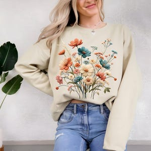 Pode incluir: Camisola bege com um design floral colorido. As flores s&atilde;o em tons de laranja, verde-azulado, rosa e creme, criando um visual vibrante e art&iacute;stico. A camisola &eacute; um estilo casual e confort&aacute;vel.