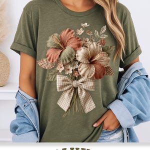 May include: Olive green t-shirt featuring a floral bouquet design. The bouquet includes hibiscus flowers in shades of peach and cream, with green leaves and a plaid bow. The shirt has rolled-up sleeves.