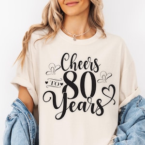 May include: Cream-colored t-shirt with the text "Cheers to 80 Years" in black script. The design includes a large number 80 and heart accents. The shirt is worn with a denim jacket.