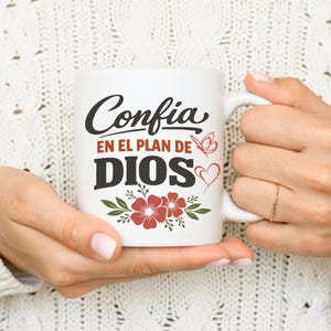 May include: White ceramic mug with the Spanish text "Confia en el Plan de Dios" in black script and bold letters. The mug features red flowers, a butterfly, and a heart design. The mug is held by a person.