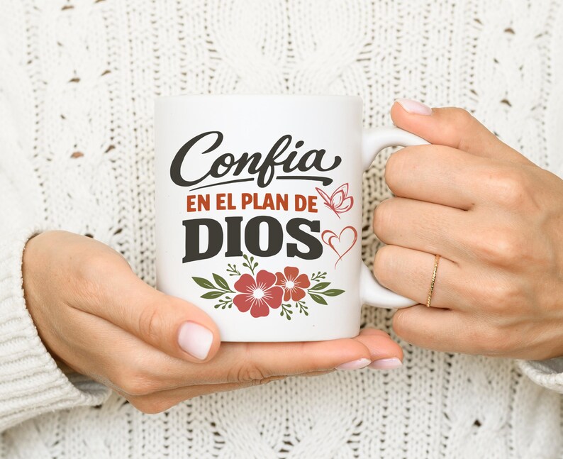 May include: White ceramic mug with the Spanish text "Confia en el Plan de Dios" in black script and bold letters. The mug features red flowers, a butterfly, and a heart design. The mug is held by a person.