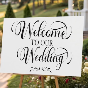 May include: A white sign with the words "Welcome to Our Wedding" in elegant black script. The sign is displayed on a wooden easel outdoors, surrounded by greenery and flowers, perfect for a wedding ceremony.