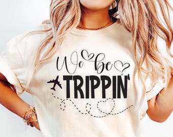 Può includere: T-shirt color crema con la scritta nera "We be TRIPPIN'" in corsivo e maiuscolo. Presenta un piccolo aereo e linee tratteggiate con cuori. Design casual a tema viaggio.