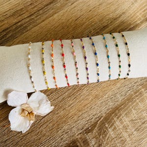 Peut inclure: Plusieurs bracelets de chaîne dorés, ornés de perles émaillées colorées. Ils sont présentés sur un rouleau de tissu blanc, avec des couleurs variées : blanc, jaune, orange, rouge, rose, violet, bleu, vert et noir.
