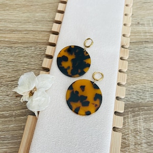 Puede incluir: Dos pares de pendientes redondos de carey con aros dorados. Los pendientes se exhiben sobre una superficie blanca con un expositor de madera y una flor blanca. Los pendientes tienen un patrón marrón oscuro y ámbar.