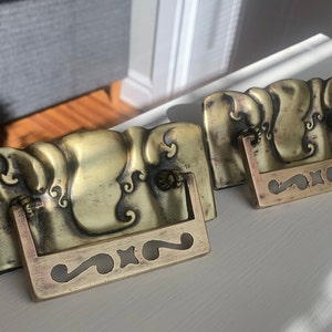 May include: Two antique brass drawer pulls with ornate designs. The pulls have a rectangular base with a decorative handle.