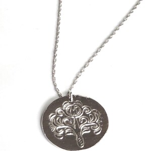 May include: A silver-toned necklace featuring a circular pendant with a stamped, symmetrical tree-like design. The pendant hangs from a thin, twisted rope chain. The metal surface has a polished finish that reflects light across the embossed pattern.