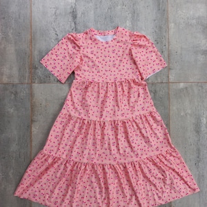 Könnte beinhalten: Ein rosafarbenes, mehrschichtiges Kleid mit Kirschendruck und kurzen Ärmeln. Das Kleid hat einen runden Ausschnitt und einen weiten Rock.