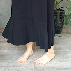 Pode incluir: Um vestido preto com bainha rodada. O vestido é feito de um tecido fluido e tem um design simples e elegante. A bainha fica a meio da panturrilha. O vestido é usado descalço num chão ladrilhado.
