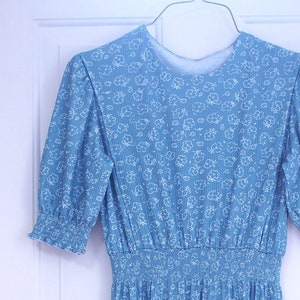 Pode incluir: Um vestido azul claro com padrão floral branco. O vestido tem um decote redondo, mangas curtas e um corpete ajustado. A saia é franzida na cintura e cai até os joelhos.