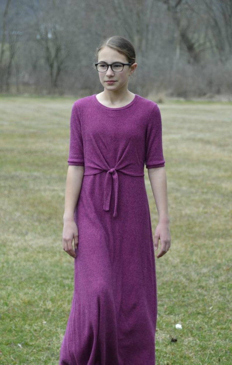 May include: A young person wearing a purple knit dress with a tie at the waist. The dress has a round neckline and short sleeves. The person is wearing black framed glasses and is standing in a grassy field.