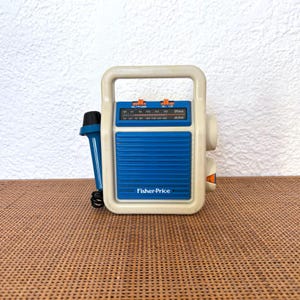 May include: A vintage Fisher-Price toy radio in cream and blue. The radio features a handle, a blue speaker panel, and a tuning dial. A blue microphone is connected. The Fisher-Price logo is visible on the front of the radio.
