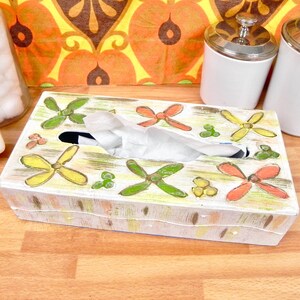 May include: A rectangular, vintage tissue box with a white base, decorated with hand-painted, colourful floral designs. The box features green, yellow, and orange flower-like shapes. White tissues are visible from the top opening.