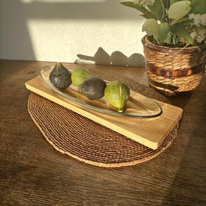 Może przedstawiać: Drewniana taca do serwowania z przezroczystą szklaną wkładką zawiera cztery dekoracyjne figi w odcieniach zieleni i ciemnego fioletu. Taca stoi na tkanej podkładce, z rośliną doniczkową w tle.