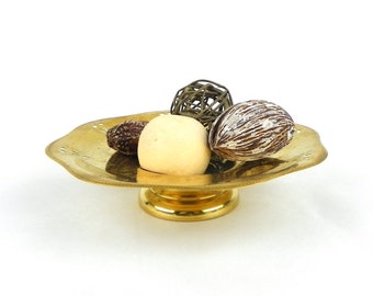 May include: A gold metal decorative bowl with a scalloped edge and a pedestal base. The bowl is filled with decorative items, including a white ball, a brown seed pod, and a woven ball.