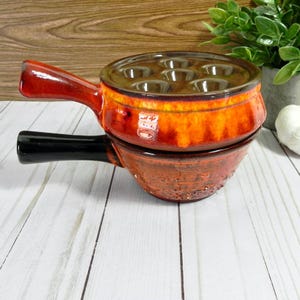 May include: Two-piece ceramic escargot dish set. The top dish is orange and brown with six indentations for snails. The bottom dish is a darker brown. Both have handles, one red and one black. The set is on a white wooden surface.