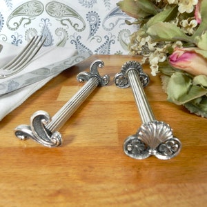 Könnte beinhalten: Zwei silberne Serviettenringe mit verzierten Designs. Die Ringe sind aus Metall und haben eine polierte Oberfläche. Sie sind beide in Form einer Säule mit dekorativen Details oben und unten.