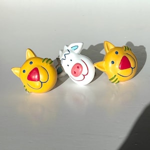 May include: Three colourful cabinet knobs. Two are yellow with red noses and green stripes, resembling cartoon cats. The third is white with a pink nose and blue accents, resembling a zebra. These decorative knobs add a playful touch.