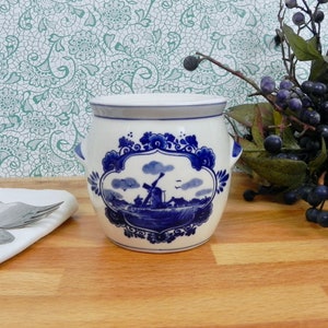 May include: A white ceramic jar with a blue and white Delft design featuring a windmill and a landscape. The jar has two handles and a lid.