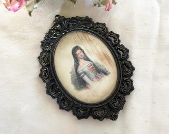 Puede incluir: Un marco de fotos ovalado de estilo antiguo con un acabado oscuro y ornamentado. El marco contiene una impresión vintage de una mujer con un vestido blanco y azul. Rosas rosas secas en el fondo.
