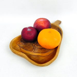 May include: A wooden, leaf-shaped bowl filled with fresh fruit. The bowl is carved from light brown wood and contains two red plums and a bright orange. The fruit is arranged in the centre of the bowl.