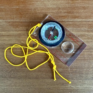 Puede incluir: Una brújula rectangular transparente con esfera negra y cordón amarillo. La brújula tiene un bisel giratorio con marcas de grados y una aguja roja. Una lupa está integrada en la base.