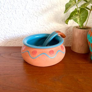May include: A small, round, terracotta-coloured bowl with a turquoise interior and rim. The bowl is decorated with pink polka dots and a wavy blue line. A matching spoon rests inside. The bowl is likely used for serving dips or sauces.