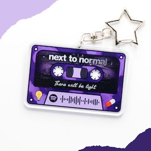 Peut inclure: Un porte-clés en acrylique transparent en forme de cassette violette avec le texte "next to normal" et "There will be light". Le porte-clés a un fermoir en forme d'étoile argentée et un porte-clés argenté.