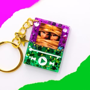 Puede incluir: Un llavero de purpurina morado y verde con una imagen de Rapunzel y Flynn Rider de la película Enredados. El llavero tiene un anillo de llave dorado y un dije de estrella.