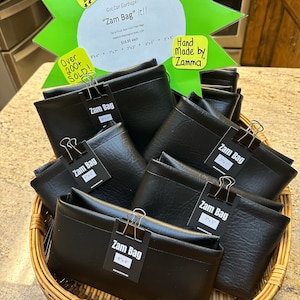 May include: A wicker basket filled with black leather car trash bags labeled "Zam Bag" in various sizes, with a green starburst sign above that reads "Got Car Garbage? "Zam Bag" it!!" and "Hand Made by "Zamma"".