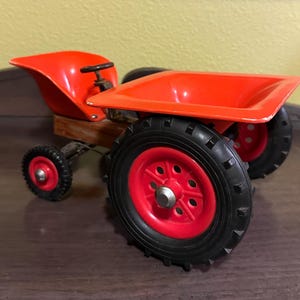 May include: A vintage toy tractor with a bright red body and wheels. The tractor features a small black steering wheel and a wooden base. The wheels have black tires with red rims. The tractor is a classic toy.