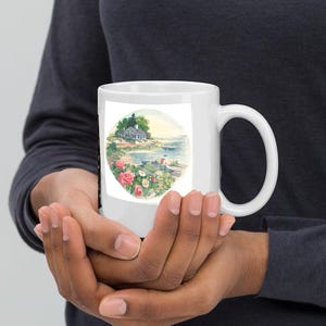 Puede incluir: Taza de cerámica blanca con una ilustración colorida de una casa junto al agua con flores rosas y árboles verdes. La taza tiene un asa.