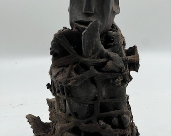 May include: A dark wooden sculpture of a human figure with a carved face. The figure is bound with rope and other materials, creating a textured and intricate design.