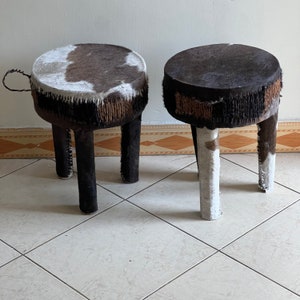 May include: Two cowhide stools with black and white patterned tops and wooden legs. The stools are on a tiled floor.