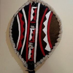 Peut inclure: Un bouclier en forme de feuille, avec un centre noir orné de motifs géométriques rouges et blancs. Le bouclier est bordé de fourrure et possède une poignée en cuir. Le design est symétrique et audacieux, d'esthétique tribale.