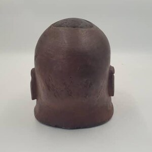 May include: A brown ceramic head-shaped planter with a textured surface. The planter has a rounded top with a small opening and two ear-like protrusions on the sides. The planter is suitable for small plants.