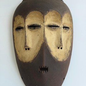 May include: A brown, oval-shaped mask with three faces. The faces are painted with light beige and brown paint. The eyes are represented by horizontal lines, and the mouth has a rectangular opening. The top of the mask has a textured design.