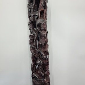 以下が含まれることがあります： 複数の図形が積み重ねられた、背の高い暗い木製の彫刻。図形は様式化された抽象的なスタイルで彫刻されており、踊ったり祝ったりしているように見える。